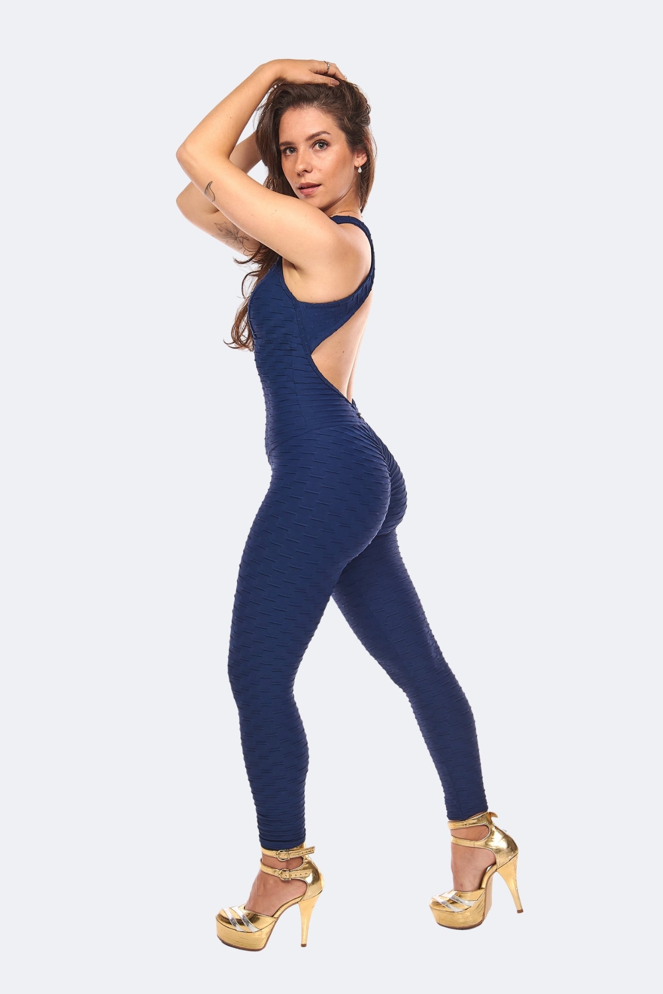 A woman modeling a navy blue ribbed one-piece jumpsuit with a back cutout, paired with gold high-heeled sandals, against a light gray background.