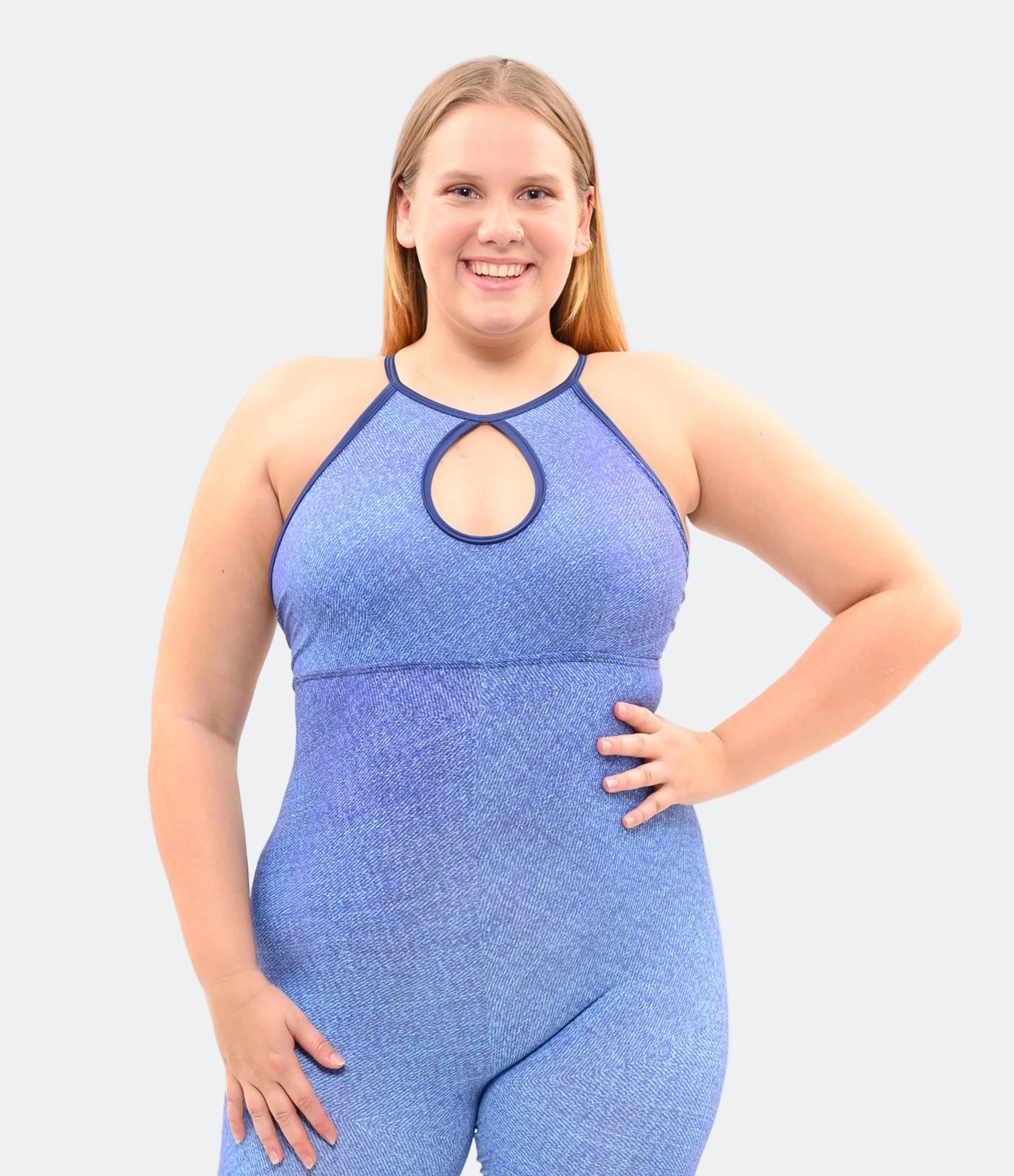 A woman standing and posing in a blue faux denim jumpsuit with a tear drop front design and adjustable lace at the back, with white platform heels.