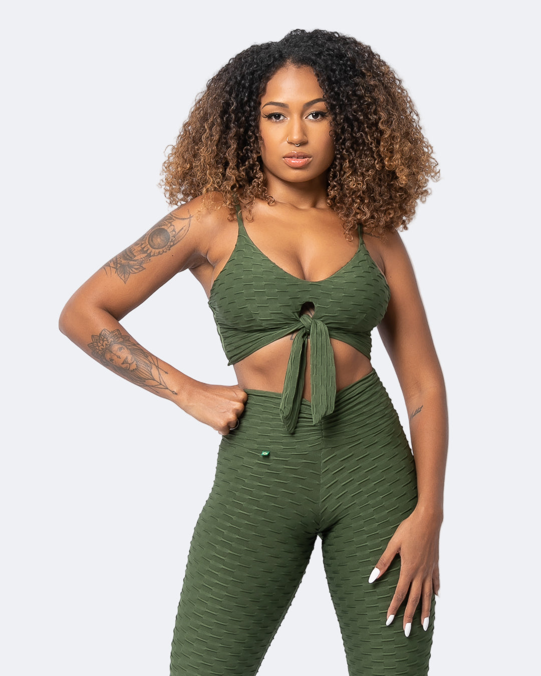 A woman wearing a tie-front sports bra in a forest green color, matching with other active wear. The sports bra has a padded top and a tie on the front.